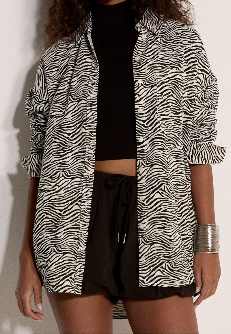 Femme portant un crop top noir, un short noir ample et une chemise rayée zèbre boutonnée avec manches retroussées et un bracelet manchette argenté au poignet droit.