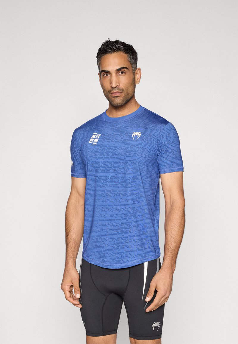 Μπλε αθλητική μπλούζα με γεωμετρικό μοτίβο, στρογγυλό λαιμόκοψη, κοντά μανίκια, συνδυασμένη με μαύρο σορτς με λευκές λεπτομέρειες και λογότυπο.