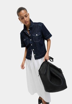 Woman wearing short-sleeve dark denim jacket over white dress, holding large black woven tote bag, looking downward against white background.