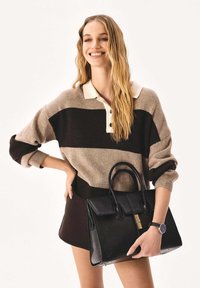 Fekete bőr kézitáska strukturált kialakítással, arany színű fém részekkel és dupla fogantyúval. A modell bézs és barna csíkos pulóvert, valamint sötét szoknyát visel.