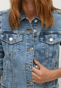 Vaaleansininen farkkutakki, jossa on hopeanväriset napit, kaksi rintataskua ja oranssit tikkaukset. Suoraleikkainen malli hieman haalistuneella tekstuurilla.