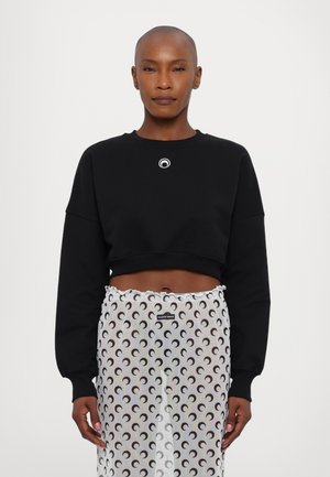 Sweatshirt noir court avec un logo rond sur la poitrine, associé à une jupe transparente présentant un motif répétitif de croissants de lune en noir et blanc.