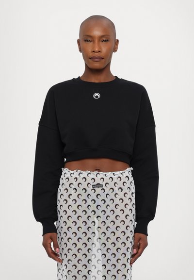 Sweatshirt noir court avec un logo rond sur la poitrine, associé à une jupe transparente présentant un motif répétitif de croissants de lune en noir et blanc.