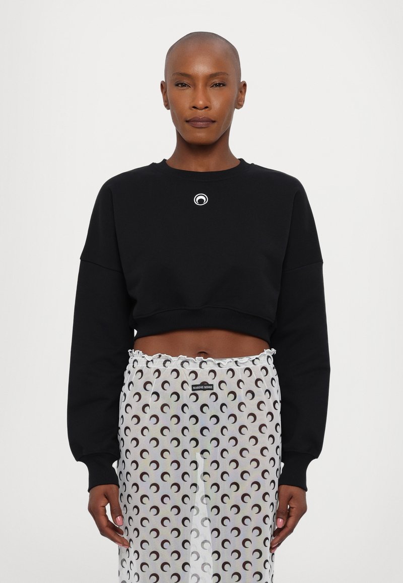 Black cropped sweatshirt with a round logo on the chest, paired with a sheer skirt featuring a repeating crescent moon pattern in black and white.