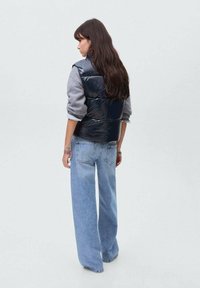 Svart pufferväst över en grå långärmad skjorta, kombinerat med ljusblå vida jeans. Bakifrån visas det släta materialet och subtila detaljer.