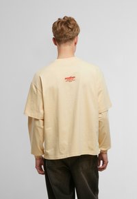 Beige, oversized, rövid ujjú póló, hátul egy kis narancssárga nyomott logóval. Pamut anyag, laza fazon.