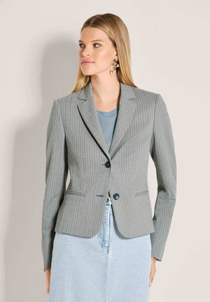 Frau mit glattem blondem Haar, die silberne, schlangenförmige Ohrringe trägt, grauen Nadelstreifenblazer über blauem Hemd und hellblauen Rock.