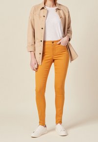 Orange skinny jeans paired with a beige denim jacket and a white t-shirt, completed with white sneakers. Smooth fabric and fitted design.