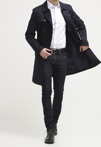 Laivastonsininen pitkä takki, jossa on kaksiriviset napit, valkoinen paita alla, tummat farkut ja mustat saappaat. Takissa on sileä pinta ja istuva leikkaus.