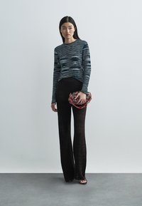 Golvend gestreept gebreid top in blauw en grijs, gecombineerd met zwarte, gestructureerde broek met wijde pijpen. Model houdt een clutch met rode accenten vast.