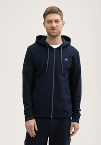 Hanorac cu fermoar de culoare bleumarin fabricat dintr-un material texturat, având glugă căptușită, buzunare frontale și șnururi de contrast. Modelul stă cu fața la cameră.