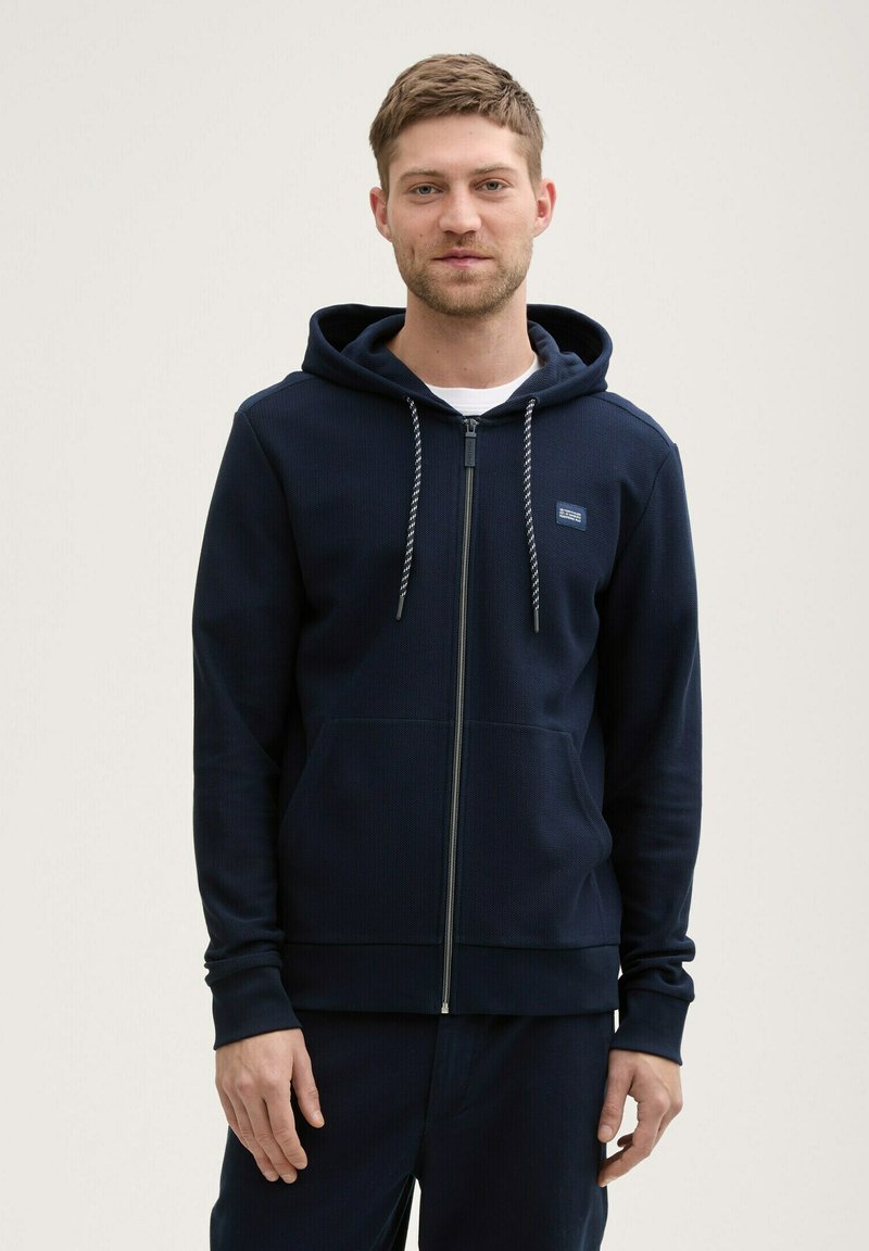 Hanorac cu fermoar de culoare bleumarin fabricat dintr-un material texturat, având glugă căptușită, buzunare frontale și șnururi de contrast. Modelul stă cu fața la cameră.