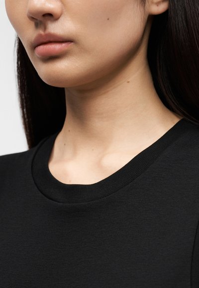 Κοντινή λήψη του κάτω μέρους του προσώπου και του λαιμού μιας γυναίκας που φοράει μαύρο μπλουζάκι με στρογγυλή λαιμόκοψη, με στενά ίσια σκουρόχρωμα μαλλιά ορατά στη μια πλευρά.