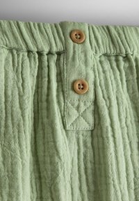 Gros plan sur un tissu vert clair texturé avec une ceinture élastique et deux petits boutons ronds en bois sur une patte de boutonnage.