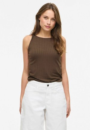 Młoda kobieta z długimi brązowymi włosami, ubrana w brązowy prążkowany top bez rękawów i białe spodnie z wysokim stanem na jednolitym tle.