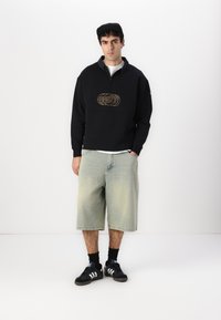 Suéter negro con cuello con cremallera y logo bordado en oro, combinado con pantalones cortos de mezclilla de color azul claro y zapatillas negras con franjas blancas.