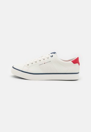 Zapatilla de cuero blanca con acentos azules, lengüeta trasera roja y diseño perforado. Presenta una textura suave y una silueta de perfil bajo.