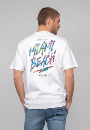 T-shirt blanc à manches courtes avec un graphique coloré "MIAMI BEACH" dans le dos, porté avec un jean bleu visible à la taille.