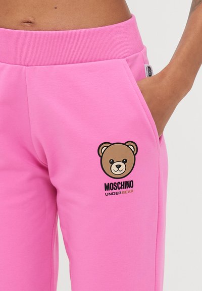 Roza trenirke iz mehke kombinacije bombaža, z grafiko medveda in logotipom "Moschino Underbear" na levi strani. Podrobnost elastičnega pasu.