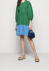 Robe colorée en vert et bleu, avec des manches bouffantes, un décolleté en forme de clé et une coupe ample, assortie à des sandales à plateforme beiges et un petit sac bleu.