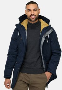 Marine parkajakke med pelsforet krave, grå detaljer, flere lynlåslommer og justerbar hætte, båret over en mørk turtleneck.
