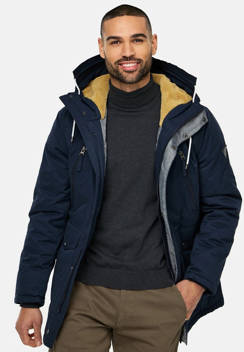 Marine parkajakke med pelsforet krave, grå detaljer, flere lynlåslommer og justerbar hætte, båret over en mørk turtleneck.