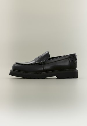Μαύρο δερμάτινο loafer με παχιά λαστιχένια σόλα, λεπτομέρεια με λουράκι penny και ραμμένο σχέδιο moc toe σε ουδέτερο φόντο.