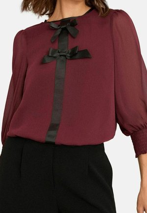Femme portant un chemisier bordeaux avec manches bouffantes transparentes, ruban vertical noir et deux nœuds noirs, assorti à un pantalon noir.