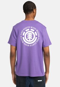 Lilla bomulds T-shirt med rund halsudskæring, prydet med et stort hvidt grafisk motiv på ryggen, der inkluderer elementære symboler og geometriske mønstre.