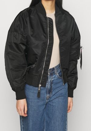 Person trägt eine schwarze Bomberjacke mit Frontreißverschluss, beige Hemd und hoch taillierte blaue Jeans, steht vor schlichtem Hintergrund.