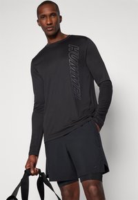 Homme en t-shirt noir à manches longues avec texte "HUMMEL" et short de sport noir tenant des bandes de résistance noires sur fond blanc.