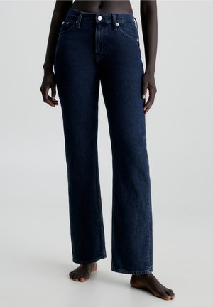 Dunkelblaue Slim-Fit-Jeans mit hoher Taille, getragen von einer dunkelhäutigen Person, die barfuß auf einem weißen Boden steht.