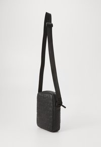Mala crna crossbody s podesivim remenom i ugraviranim uzorkom logotipa "HUGO" na prednjoj strani, prikazana na jednostavnoj pozadini.