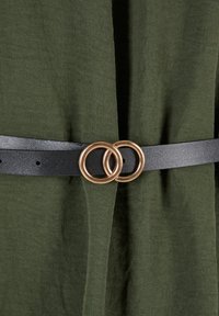 Zwarte leren riem met twee met elkaar verbonden gouden ronde gespen, gedragen over olijfgroene stof.