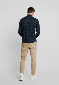 Camicia in flanella a scacchi verde scuro e blu navy, con colletto classico e dettagli dei bottoni. Abbinata a chinos beige e sneakers bianche.