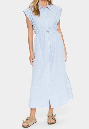 Kobieta ubrana w sukienkę midi bez rękawów w pionowe, jasnoniebiesko-białe paski, z guzikami z przodu i wiązanym paskiem w talii, w połączeniu z brązowymi sandałami na klamrę.