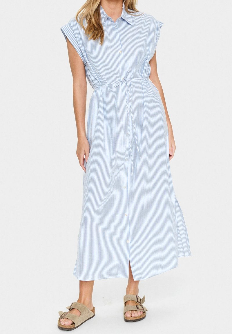 Femme portant une robe midi sans manches à rayures verticales bleu clair et blanc, boutonnée à l'avant avec une ceinture nouée à la taille, associée à des sandales marron à boucle.