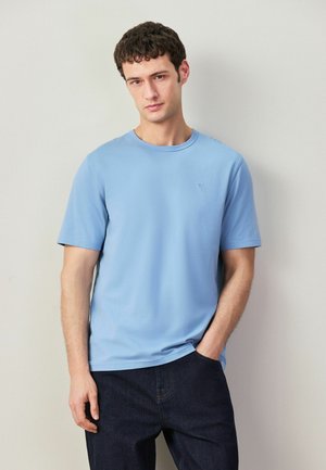 Young man wearing a light blue short-sleeve t-shirt and dark jeans, standing with one hand in pocket against a neutral background.