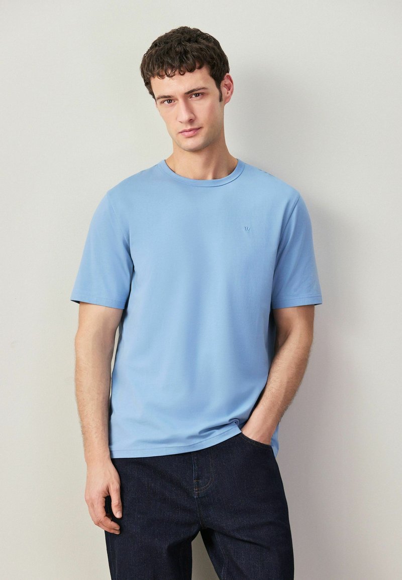 Mladý muž v svetlomodrom tričku s krátkym rukávom a tmavých džínsoch, stojaci s jednou rukou v vrecku na neutrálom pozadí.