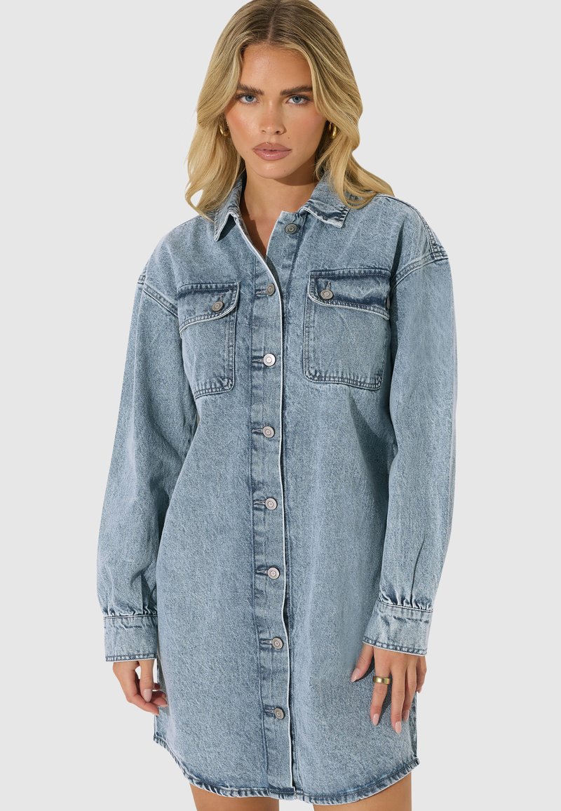 Mujer rubia que lleva un vestido camisa vaquero azul claro con botones, mangas largas y bolsillos delanteros en el pecho, de pie sobre fondo liso.