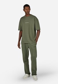 Ελιάς πράσινη υπερμεγέθη μπλουζάκι και ταιριαστά παντελόνια, κατασκευασμένα από μαλακό ύφασμα, με λογότυπα. Το μοντέλο φοράει λευκά αθλητικά.