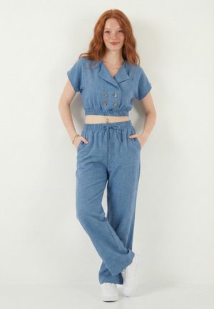 Roodharige vrouw staat met handen in de zakken, draagt een blauw crop top met knopen en bijpassende losse broek, gecombineerd met witte sneakers.