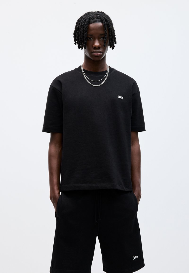Sort bomulds T-shirt med korte ærmer og rund hals, med et lille hvidt logo på brystet. Sammen med sorte shorts med snørre.