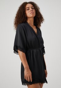Robe en satin noir avec des manches courtes et larges, et des bordures en dentelle aux poignets et à l'ourlet. Présente un design enroulé avec une ceinture assortie à la taille.