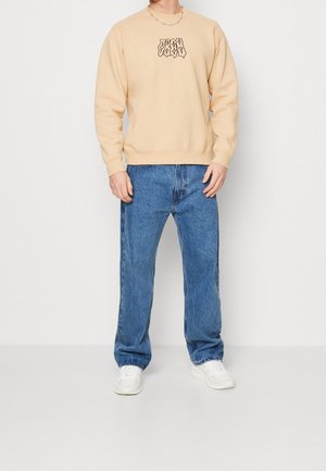 Sudadera beige con texto gráfico negro, combinada con pantalones de mezclilla azules y zapatillas blancas. Conjunto casual de corte relajado.
