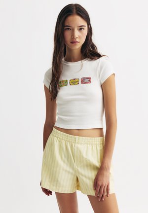 Witte cropped t-shirt met drie kleurrijke illustraties van ingeblikt voedsel op de voorkant, gecombineerd met gele gestreepte elastische shorts.