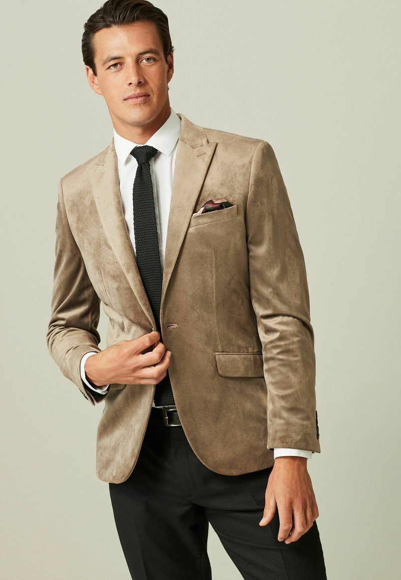 Beige Wildleder-Blazer mit Revers, zwei Fronttaschen und einem dekorativen Einstecktuch. Kombiniert mit einem weißen Hemd und einer schwarzen Krawatte.