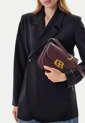 Woman in black blazer holding a dark brown leather handbag with gold-tone clasp and adjustable strap across her chest.