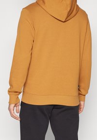 Sudadera con capucha marrón de acabado texturizado, puños y dobladillo acanalados, combinada con pantalones oscuros, mostrando un diseño casual y cómodo.