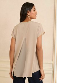 Blusa beige con maniche corte, orlo rotondo sul retro con una fessura centrale e una texture liscia, abbinata a jeans scuri.